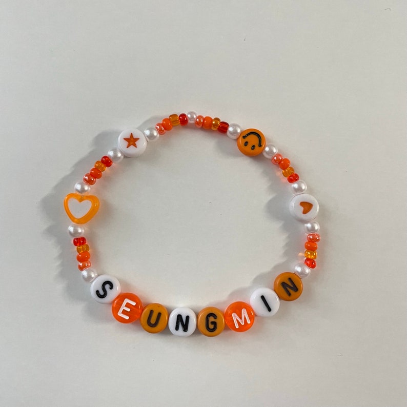 May include: Orange and white beaded bracelet with the name "SEUNGMIN" spelled out in white beads. The bracelet also features a star, heart, and smiley face charm.