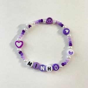 May include: Purple and white beaded bracelet with the name "MINHO" spelled out in white letters with black outlines. The bracelet also features purple heart and crescent moon charms.