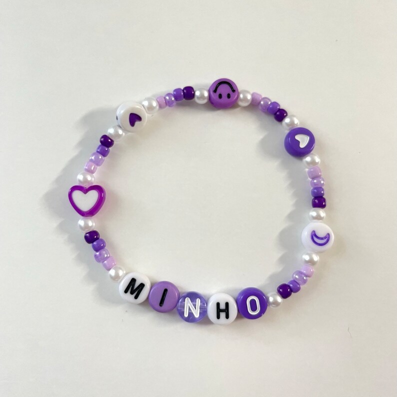 May include: Purple and white beaded bracelet with the name "MINHO" spelled out in white letters with black outlines. The bracelet also features purple heart and crescent moon charms.