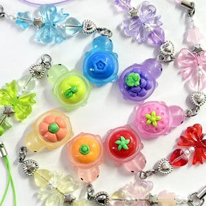 May include: A collection of colorful phone charms with fruit and flower designs. The charms feature small turtle shapes with various fruits like strawberries, oranges, and blueberries. Each charm has a matching colored cord and silver-toned hardware.
