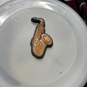 Op de afbeelding: Een bruine saxofoonbedel met zwarte rand. De muziekinstrumentbedel is gedetailleerd met gegraveerde lijnen en cirkelvormige accenten. De bedel staat tegen een lichtgrijze achtergrond.