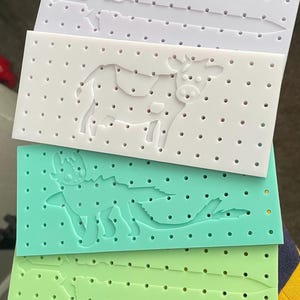 Może przedstawiać: Cztery białe tablice z otworami z różnymi wzorami. Górna tablica ma wzór miecza, druga tablica ma wzór krowy, trzecia tablica ma wzór smoka, a dolna tablica ma wzór miecza.