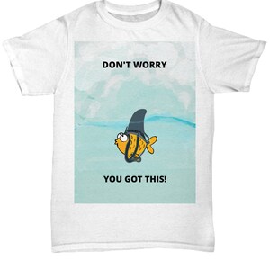 Może przedstawiać: Biały t-shirt z grafiką przedstawiającą złotą rybkę w płetwie rekina. Na koszulce nadrukowany jest tekst "Don't Worry You Got This!"