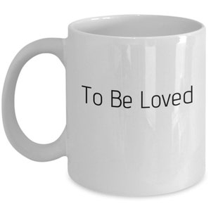 Peut inclure: Une tasse à café en céramique blanche avec une inscription noire qui dit "To Be Loved".