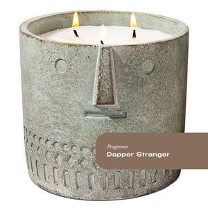 May include: A three-wick candle in a grey, face-shaped concrete container. The candle has white wax and the container has a textured surface with a nose, eyes, and mouth design. The text "Dapper Stranger" is visible.