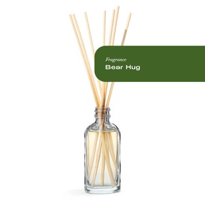 Puede incluir: Una botella de vidrio transparente con palitos de madera insertados. La botella contiene un líquido amarillo claro. La botella está etiquetada con "Fragrance Bear Hug".