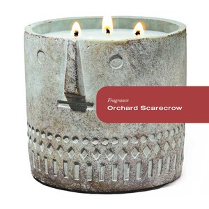 May include: A green concrete candle with a face design and a decorative pattern around the bottom. The candle has three wicks and is labelled "Orchard Scarecrow" with the word "Fragrance" above it.