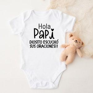 May include: White baby onesie with black text that reads "Hola Papi Diosito escuchó sus oraciones!!"  A small heart is above the word "Papi".