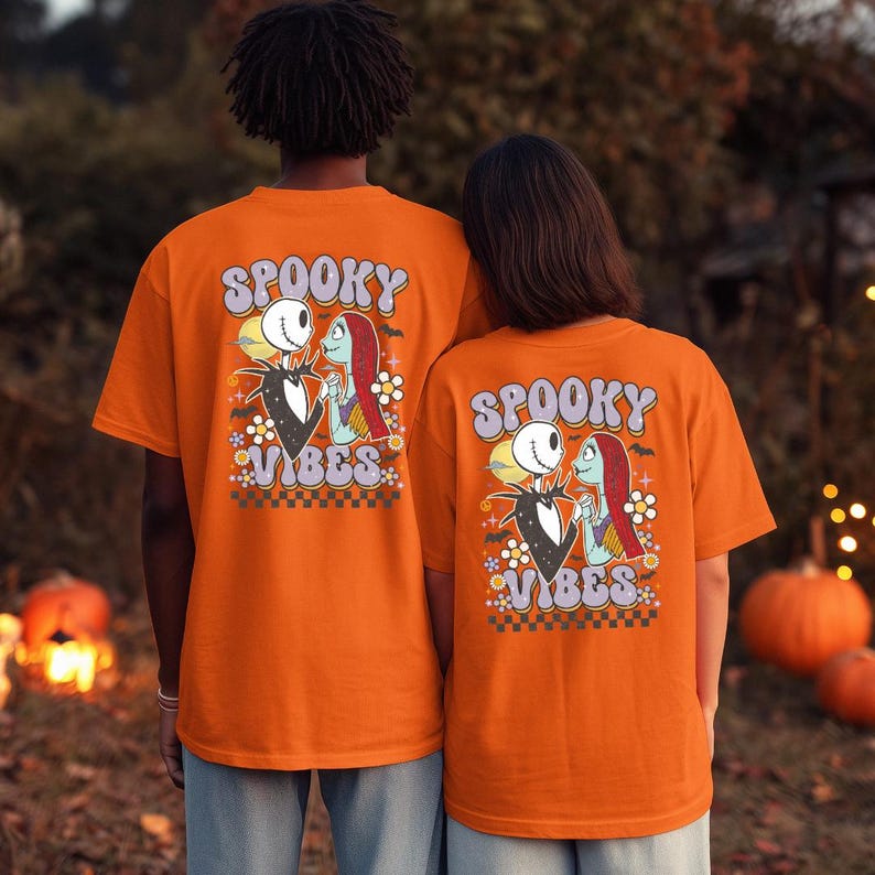 Puede incluir: Dos personas con camisetas naranjas con un gr&aacute;fico de Halloween. Las camisetas presentan una imagen de dibujos animados de Jack Skellington y Sally, con las palabras "Spooky Vibes" en una fuente retro. El fondo est&aacute; borroso, con calabazas y luces.