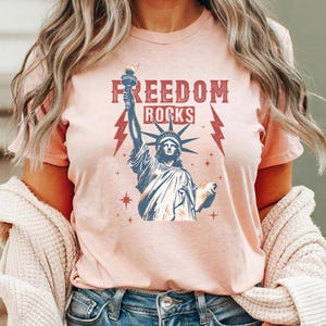 May include: A light pink t-shirt featuring a graphic of the Statue of Liberty holding a torch, with the words "FREEDOM ROCKS" in a distressed font. Red lightning bolts flank the statue. The shirt is a crew neck.