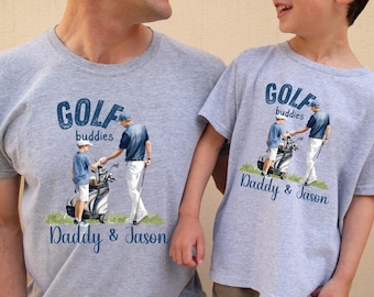 May include: Two grey t-shirts with a golf theme. The shirts feature a cartoon illustration of two golfers with golf clubs and a golf bag. The text on the shirts reads "Golf Buddies" and "Daddy & Jason".
