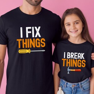May include: Black t-shirts with white and orange text. One shirt says "I FIX THINGS" with a yellow and silver screwdriver graphic. The other shirt says "I BREAK THINGS" with a silver and red hammer graphic.