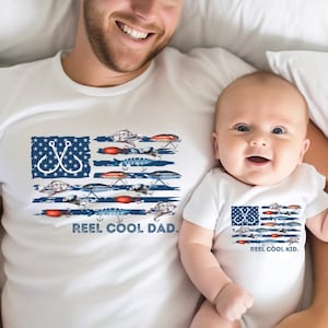 May include: A white t-shirt with a distressed American flag design featuring fishing lures and the text "Reel Cool Dad." A white baby onesie with the same design and the text "Reel Cool Kid."