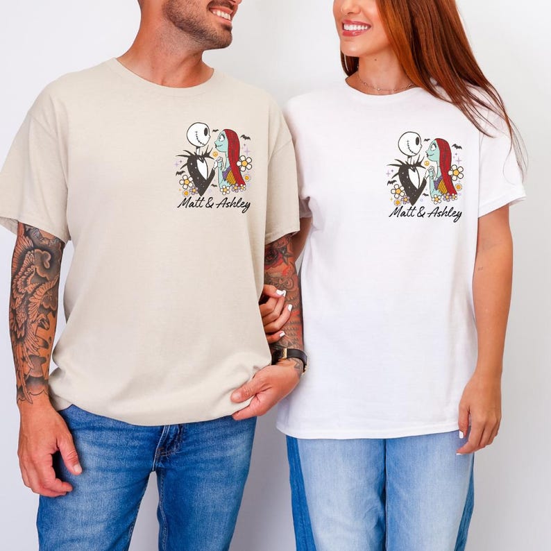 Puede incluir: Dos camisetas, una beige y otra blanca, cada una con un gr&aacute;fico de Jack y Sally de Pesadilla antes de Navidad. El gr&aacute;fico incluye el texto "Matt & Ashley" en una fuente cursiva. Ambas camisetas son de manga corta.