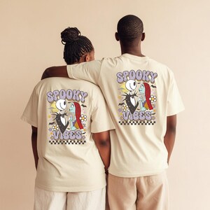 Puede incluir: Dos personas con camisetas beige con un gr&aacute;fico colorido en la espalda. El gr&aacute;fico presenta a Jack Skellington y Sally de The Nightmare Before Christmas, con las palabras "Spooky Vibes" en una fuente retro. El dise&ntilde;o incluye flores y un borde a cuadros.