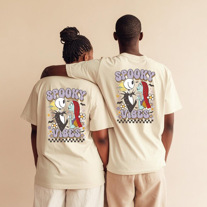 Puede incluir: Dos personas con camisetas beige con un gr&aacute;fico colorido en la espalda. El gr&aacute;fico presenta a Jack Skellington y Sally de The Nightmare Before Christmas, con las palabras "Spooky Vibes" en una fuente retro. El dise&ntilde;o incluye flores y un borde a cuadros.