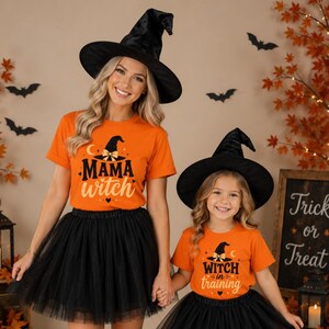 May include: Two orange t-shirts with black witch hats and text. One shirt reads MAMA witch with a hat graphic, the other reads WITCH in training with a hat graphic. Both are paired with black tulle skirts and black pointed witch hats.