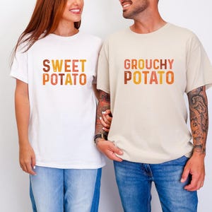 May include: Two matching t-shirts, one white and one beige, with the words "SWEET POTATO" and "GROUCHY POTATO" in a gradient orange and brown font. The shirts are worn by two people, with the white shirt on the left and the beige shirt on the right.