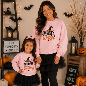 May include: Two light pink long-sleeve sweatshirts featuring black and orange graphic prints. The adult sweatshirt displays the text MAMA WITCH with a witch hat, while the child sweatshirt displays the text WITCH in training with a witch hat.