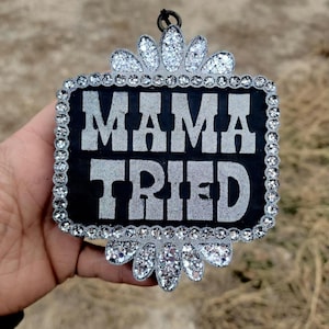 May include: A black rectangular ornament with the words "MAMA TRIED" in silver glitter lettering. The ornament is framed with a row of clear rhinestones and has decorative glitter accents at the top and bottom.