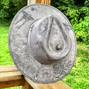 May include: A gray suede fedora hat with a floral and moth design. The hat has a crescent moon design on the crown.