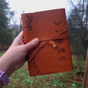 May include: A brown leather journal with a wraparound strap closure. The cover features an embossed rabbit and crescent moon design. The journal is held in a hand, showcasing the rich color and texture of the leather.