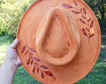 May include: An orange suede hat with a wide brim and a teardrop-shaped crown. The hat features brown leaf designs on the brim and crown. The hat is held by a person's hand against a background of green grass and trees.