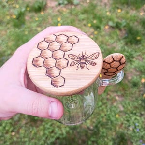 May include: A clear glass honey pot with a wooden lid and spoon. The lid features a burned-in design of a honeycomb and a bee. The spoon has a honeycomb design on the spoon head. The honey pot is held in a hand against a blurred green background.