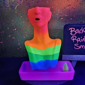 Puede incluir: Un quemador de incienso con forma de busto de colores arcoíris, con una base rosa y un pequeño cono de incienso. El quemador es en tonos naranja, verde y azul. Un letrero de pizarra dice "Backflow Rainbow + Smoke" con dimensiones de 17,8x12,7x7,6 cm.