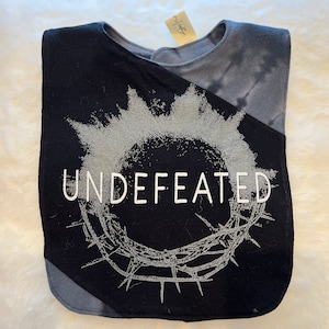 Puede incluir: Babero para bebé negro y gris con la palabra "UNDEFEATED" en letras blancas. El babero presenta un diseño de corona de espinas gris y un patrón tie-dye en la parte superior derecha. Una pequeña etiqueta está adjunta al babero.