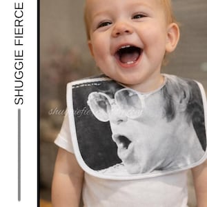 May include: A white baby bib featuring a black and white image of a person wearing sunglasses and singing. The bib is worn by a smiling baby wearing a white onesie. The text "SHUGGIE FIERCE BIB" is on the left side of the image.