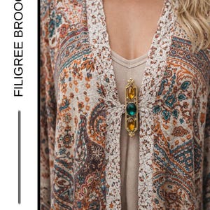 May include: A gold filigree brooch with three gemstones, orange, green, and yellow, is pinned to a patterned cardigan. The cardigan features a paisley design in shades of orange, blue, and cream. The text "FILIGREE BROOCH" is visible.