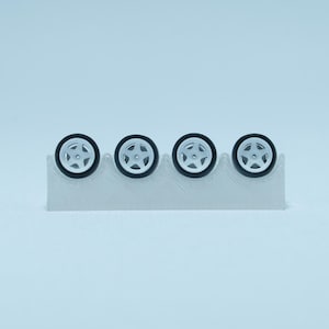 May include: Four miniature toy car wheels with white five-spoke rims and black rubber tires, arranged in a horizontal row and held in place by a light gray, textured, wave-shaped plastic display stand against a solid light blue background.