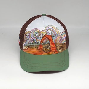 May include: A brown and green trucker hat with a white crown featuring a colorful illustration of Delicate Arch in Arches National Park, Utah.