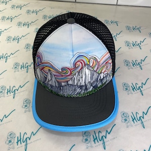 May include: A black and blue trucker hat with a white crown featuring a colorful mountain scene with a rainbow sky.