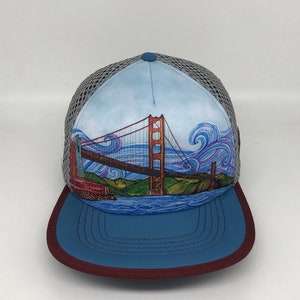 May include: A blue and gray trucker hat with a colorful illustration of the Golden Gate Bridge in San Francisco. The bridge is painted in orange and red, with a blue sky and white clouds in the background. The hat has a blue brim with a maroon edge.