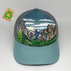 May include: A trucker hat with a scenic landscape design. The hat features a light blue brim and a gray mesh back. The front panel displays a watercolor painting of mountains, trees, and a waterfall under a blue sky with clouds and a setting sun.