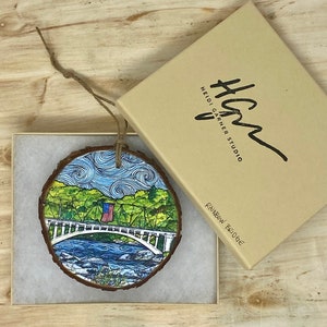 May include: A wooden ornament featuring a hand-painted illustration of a white bridge with a small American flag, surrounded by green trees and a blue river. The ornament is in a box with the text "Heidi Garner Studio" and "Rainbow Bridge".