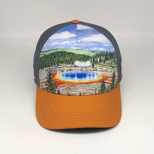 May include: A gray and orange trucker hat with a colorful illustration of a geyser in a national park. The geyser is surrounded by trees and a blue lake.