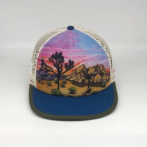 May include: A white and blue trucker hat with a colorful sunset and Joshua Tree landscape design. The hat has a blue mesh back and a green brim.