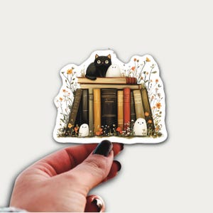 May include: A sticker depicting a black cat and a white ghost on a pile of books. The books are in various colours, and the scene includes flowers and small ghosts. The sticker is held by a hand.