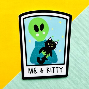 May include: A green alien with a blue sweater is holding a black cat with yellow eyes and antennas. The image is in a white frame with the text "ME & KITTY" at the bottom.