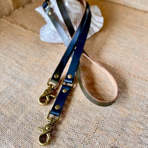 May include: Two leather dog leashes with brass hardware. One leash is black and the other is olive green.