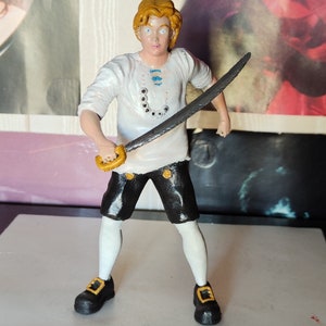 May include: A hand-painted figurine of a person wearing a white shirt with a blue and white design, black shorts, white socks, and black shoes. The figurine is holding a sword in their right hand.