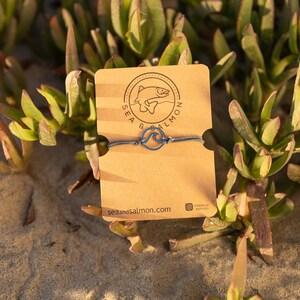May include: A blue and white string bracelet with a wave charm. The bracelet is on a brown card with the text "Sea & Salmon" and the website address "seasandsalmon.com".