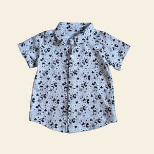 Op de afbeelding: Wit overhemd met knopen en een patroon van zwarte Mickey Mouse-figuren en stippen. Het shirt met korte mouwen heeft een kraag en is dichtgeknoopt. Het shirt is waarschijnlijk voor een kind.