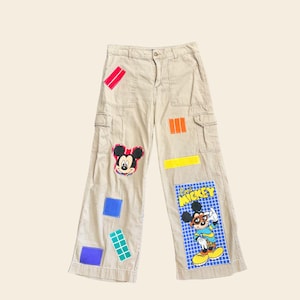 May include: Khaki cargo pants with colorful patches and cartoon graphics. Patches include Mickey Mouse and geometric shapes in red, orange, blue, green, and yellow. The pants have multiple pockets and a button closure.