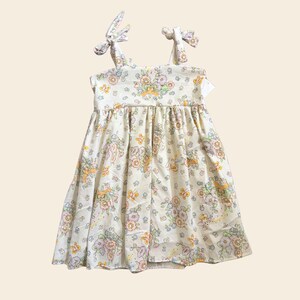 May include: A sleeveless dress in a cream-colored fabric featuring a repeating pattern of pastel-toned flowers, small butterflies, and rabbits. The garment has a gathered skirt and shoulder straps that tie into bows at the top of the shoulders.