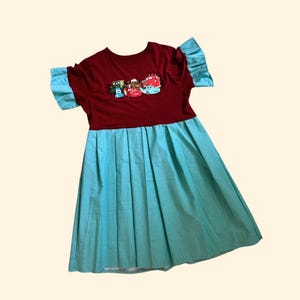 Può includere: Un vestito color amaranto con gonna a pieghe azzurra e maniche corte. La parte superiore del vestito presenta una grafica colorata di auto dei cartoni animati. Le maniche sono azzurre con un design a balze.