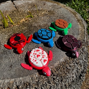 May include: Five colourful turtle figurines with different patterns on their shells. The turtles are made of plastic and are about 7.5 cm in length. The turtles are arranged in a circle on a wooden surface.